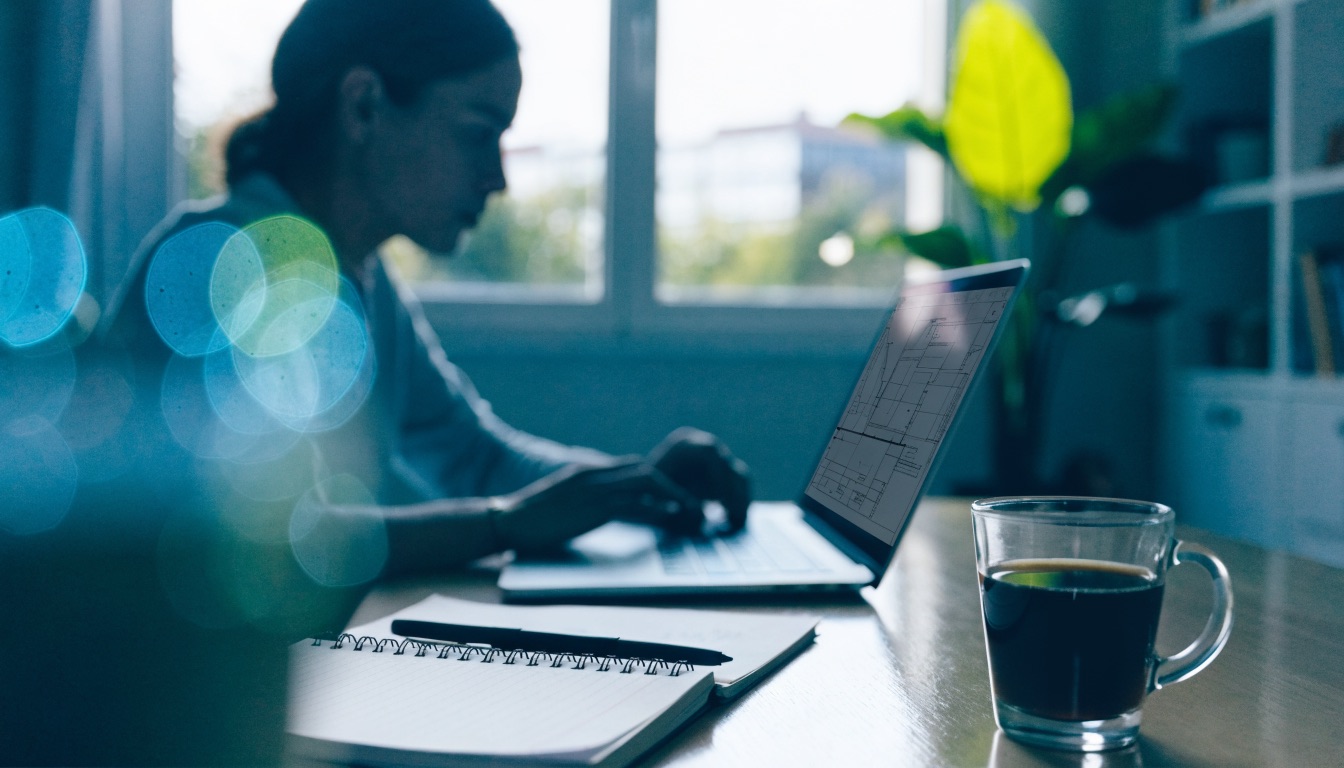 A person working at a laptop by a window, with a notebook and pen on the desk beside them